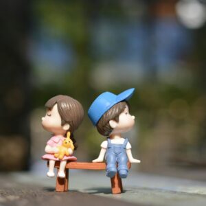 Adorable figurines of a boy and girl sit on a bench, bathed in summer sunlight.